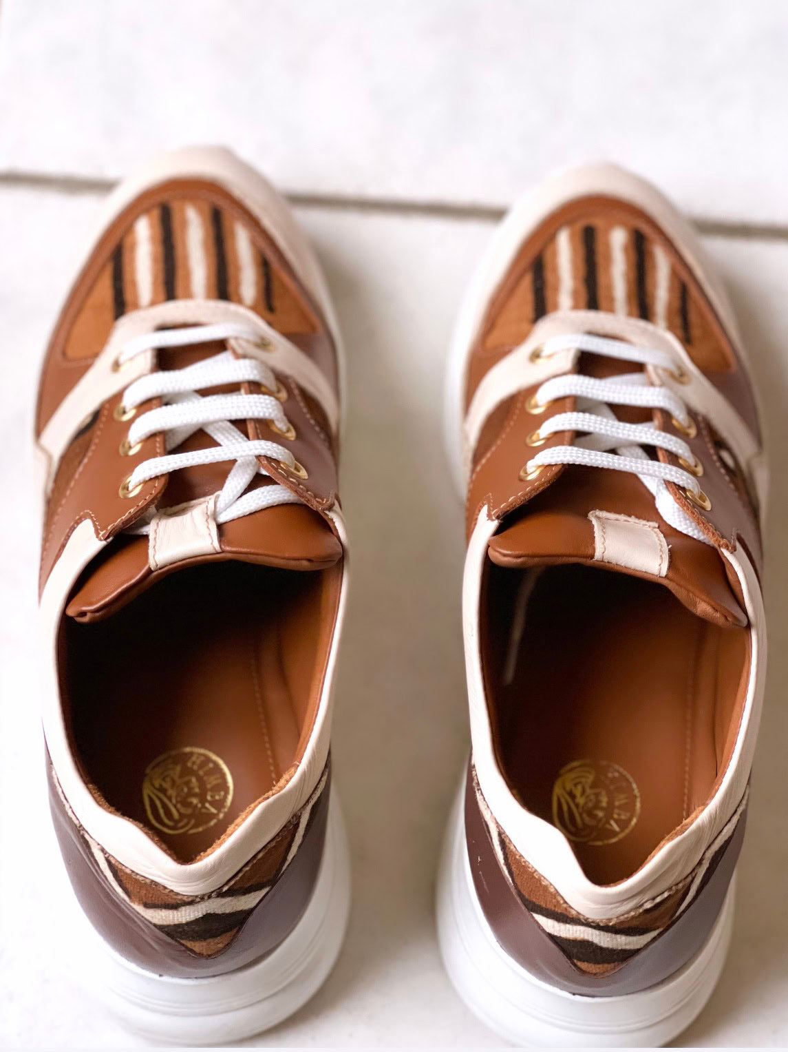 Caractéristiques: * Tige: En cuir véritable et tissu bogolan * Semelle intérieure: Confort orthopédique * Semelle extérieure: Caoutchouc robuste pour une adhérence optimale * Fermeture: Lacets en coton renforcé * Couleur: Mélange de blanc, beige et marron * Taille disponible: Du 36 au 45 Avantages: * Confort: Conception ergonomique pour un usage quotidien * Durabilité: Matériaux de haute qualité pour une longue durée de vie * Style: Design contemporain avec une touche culturelle unique * Polyvalence: Idéale